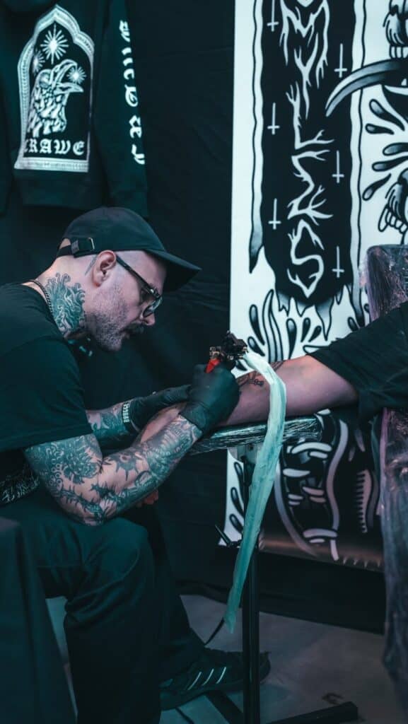 A man with a hat and glasses tattooing and arm
